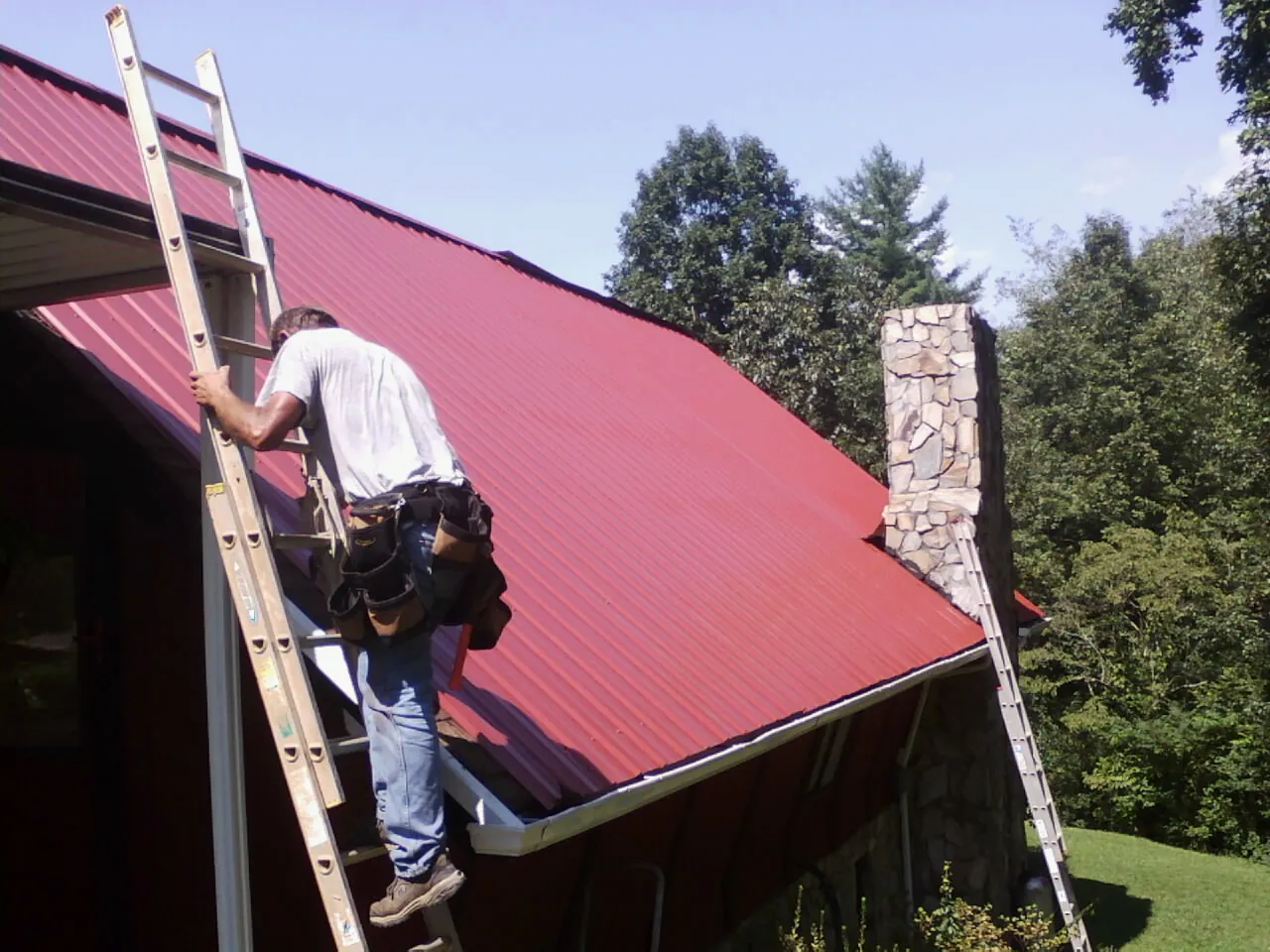 Roofer on a ladder beside a red metal roof and stone chimney — Soffit & Fascia Repair by Roofing Brothers in Kahoka